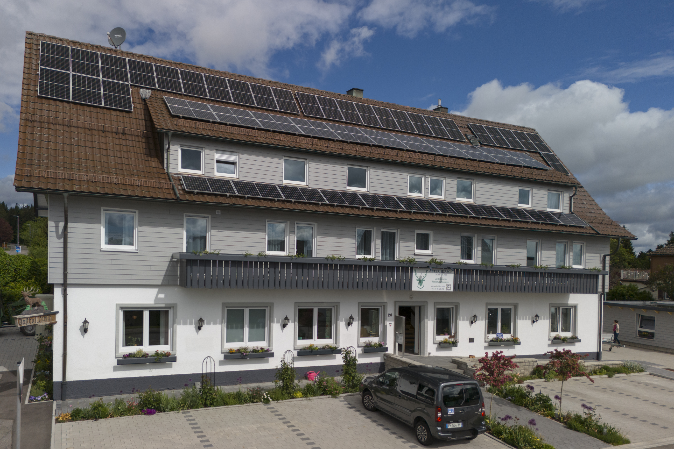 Rollstuhl-Urlaub: Außenansicht eines modernisierten Gebäudes mit großer Photovoltaikanlage auf dem Dach. Vor dem Haus befinden sich Parkplätze, ein gepflegter Eingangsbereich und Blumenbeete. Die nachhaltige Energieversorgung unterstreicht den modernen Charakter der Unterkunft. - Pension Alter Hirsch - Ferienwohnung Tanne