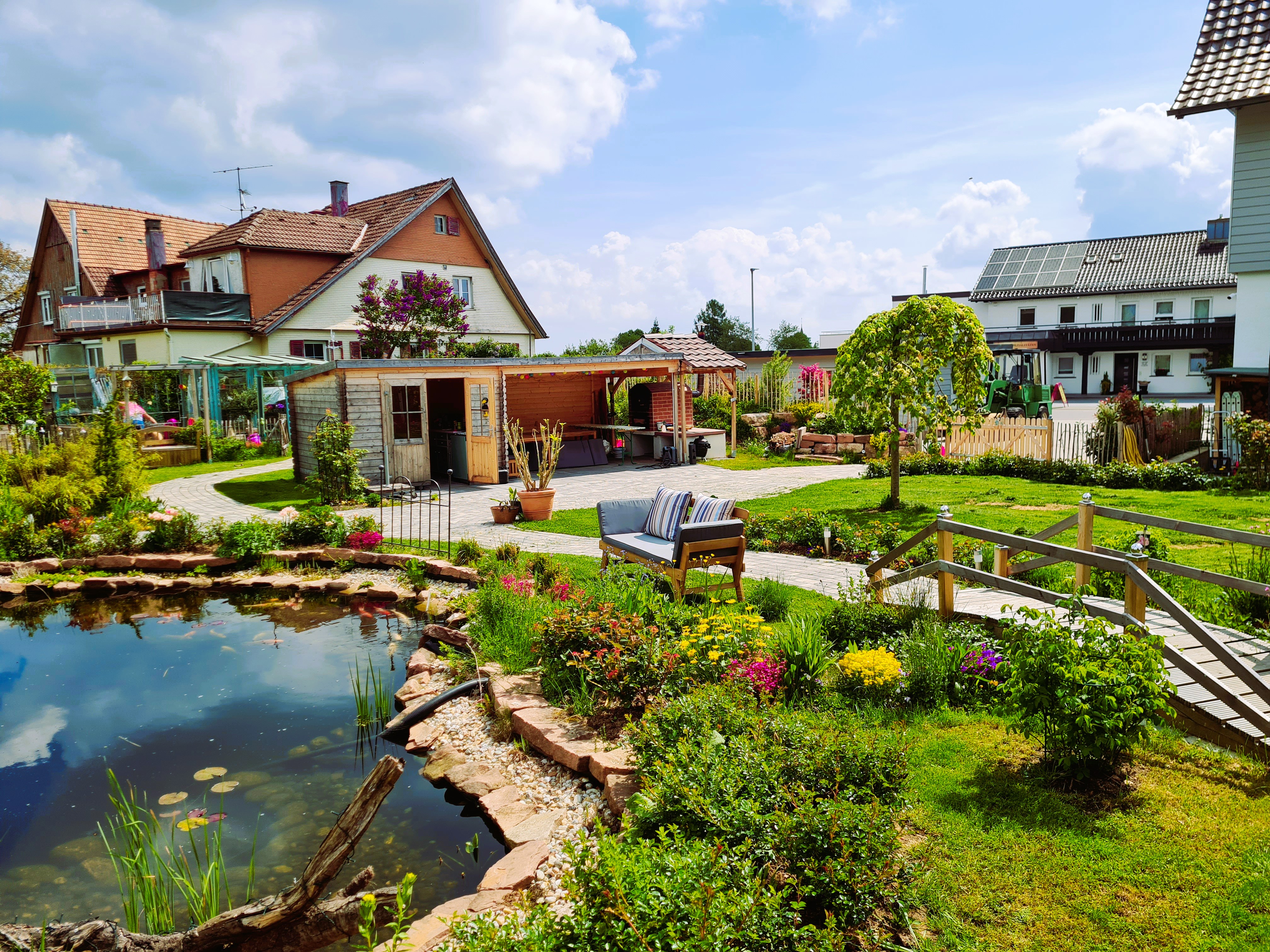 Rollstuhl-Urlaub: Großzügiger Gartenbereich mit Teich, Sitzmöglichkeiten und gepflegten Pflanzen. Im Hintergrund befinden sich weitere Gebäude und Gartenbereiche. Ein idyllischer Ort zum Entspannen im Freien. - Pension Alter Hirsch - Ferienwohnung Tanne