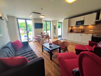 Rollstuhlgerechte Unterkunft - Unterkunftsart: Ferienhaus - offene Küche und Wohnzimmer mit 3-SitzerSofa, 2 Sesseln, 2 Hocker und 2 kleine Rattansessel - Erdhausapartment WEST